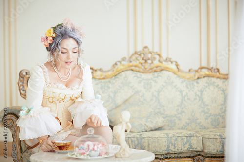 greyhead woman in Rococo dress posing in front of pink background while drinking tea photo