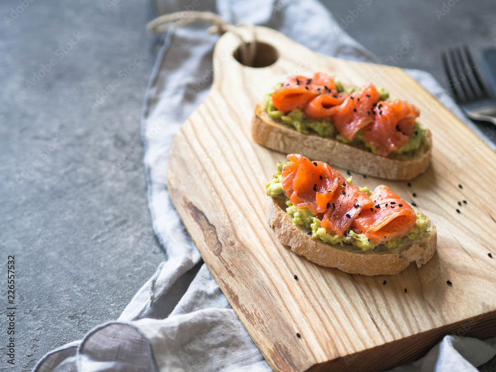 Toasts with avocado and salted salmon and black sesame seeds on a wood board on a gray background