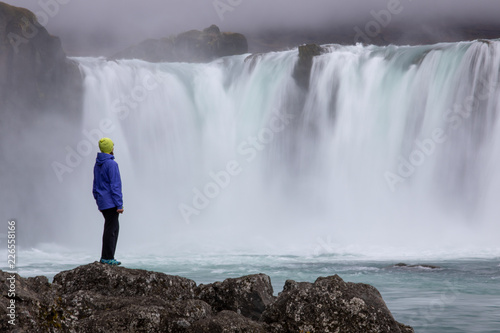 A beautiful young woman is standing against the backdrop of a beautiful waterfall. Iceland