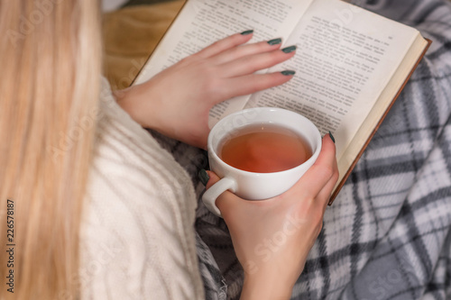 Woman holds hot cup of tea and reading book in bed. Girl wears woolen sweater and covered legs with retro blanket. Winter and cold weather concept. Close up, selective focus