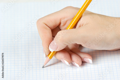 Pencil in the right female hand. Under the hand is a copybook in a cage. Close-up. Light background.