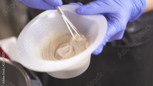 A close up of hands in blue rubber gloves holding a bowl with hair-dye and whisking it in circles, anticlockwise first, then clockwise