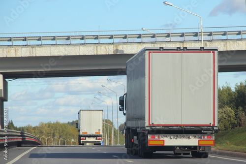 Road transport - white trucks with semi-trailers go under the bridge drive the suburban asphalt highway on a summer day against the blue sky - export-import transportation in Europe