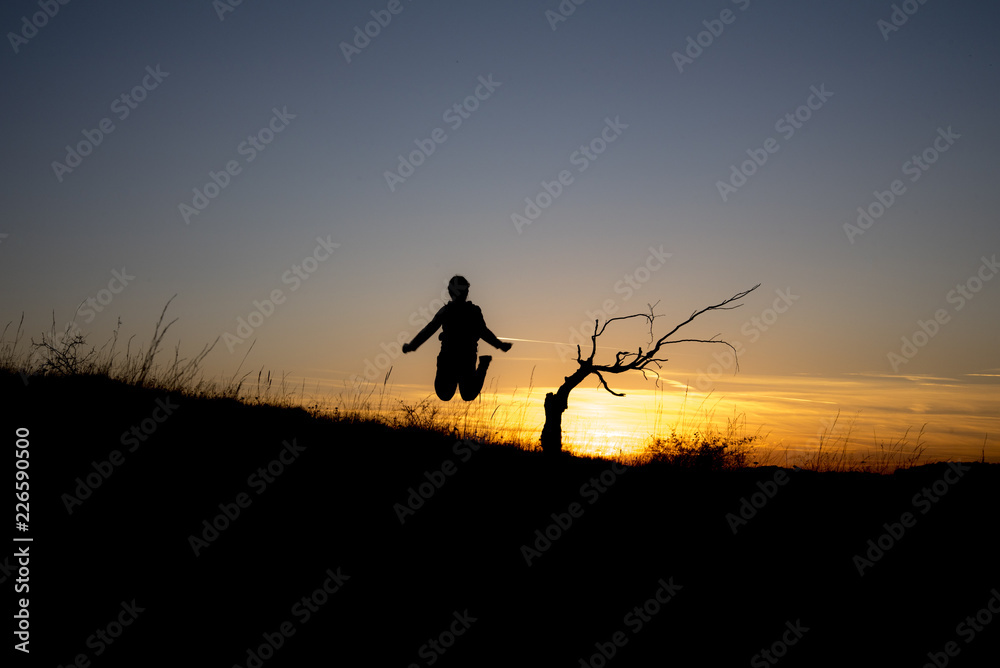 A dry tree on a hill at sunset at the beginning of October and a child jumping into the air