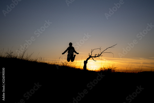 A dry tree on a hill at sunset at the beginning of October and a child jumping into the air