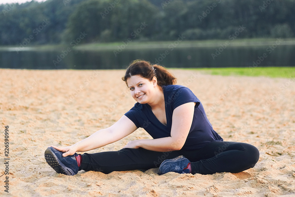 Pretty woman working out and stretching legs while sitting on sand beach near lake.