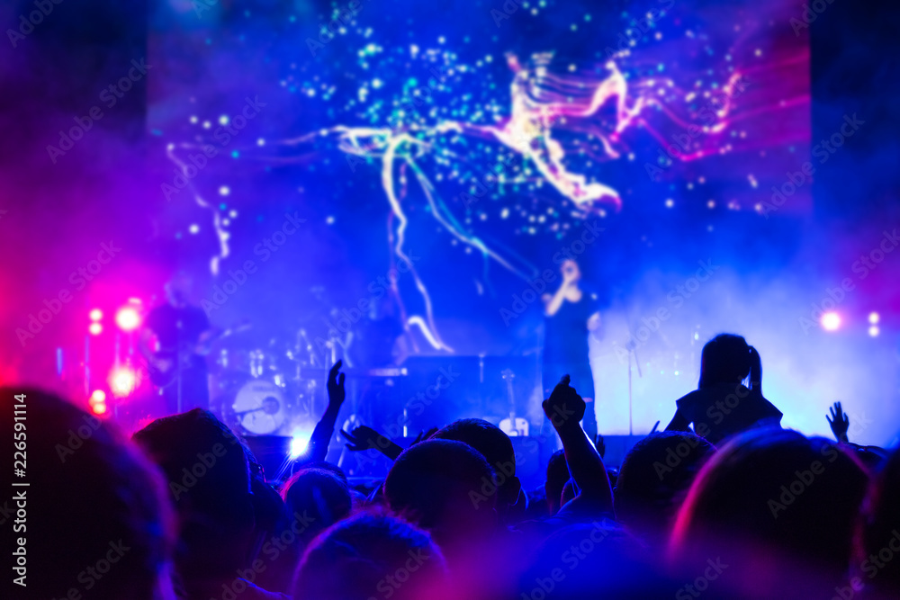 Crowd at concert. People silhouettes on backlit by bright blue and purple stage lights. Cheering crowd in colorful stage lights. Raised hands and smartphones against scene