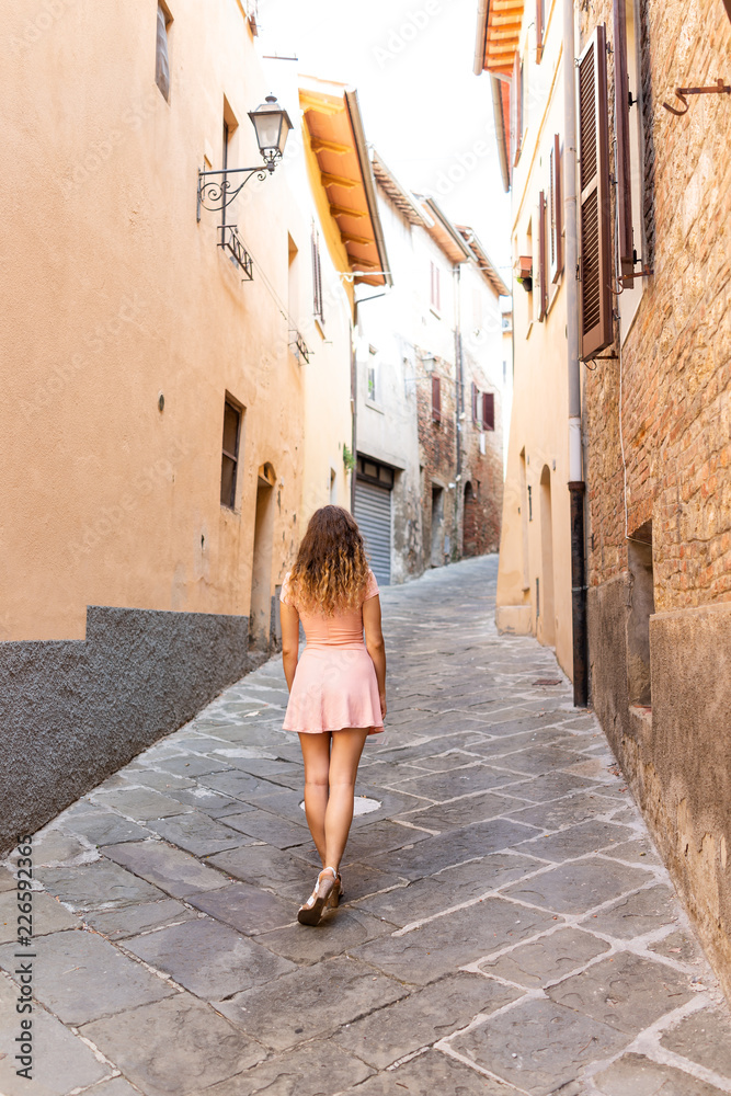Chiusi, Italy street alley in small historic medieval town village in Umbria near Tuscany during sunny summer day, back of young woman tourist walking in pink dress