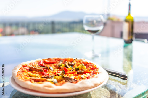 Macro closeup of fresh crust handmade pizza on glass table on terrace outside in Italy with red tomato sauce, vegetables, glass and bottle of red wine