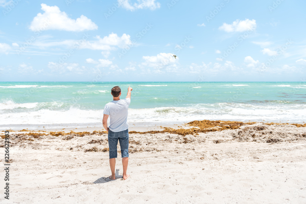 Young man in tshirt, shorts standing on sand, sandy beach in Miami, Florida with ocean, sea waves pointing finger, hand to flying bird, pelican