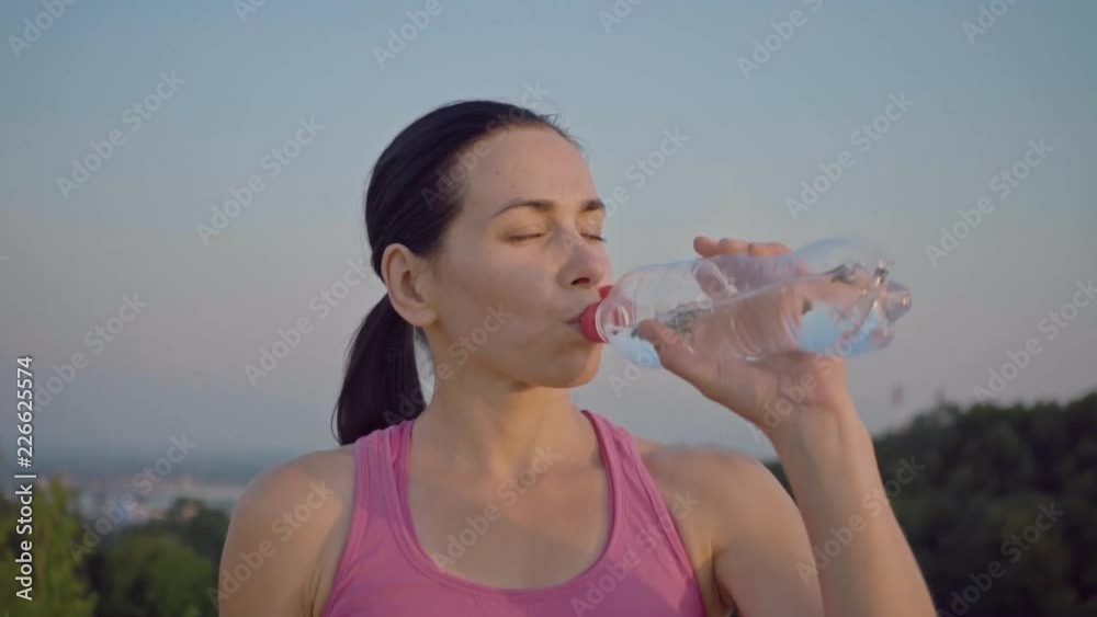 Portrait of an attractive girl on the background of the city. Sportswoman in a purple t-shirt drinks water from a bottle. Cute young woman with a sporty build is looking at the camera. Long-haired Portrait of an attractive girl on the background of the city. Sportswoman in a purple t-shirt drinks water from a bottle. Cute young woman with a sporty build is looking at the camera. Long-haired