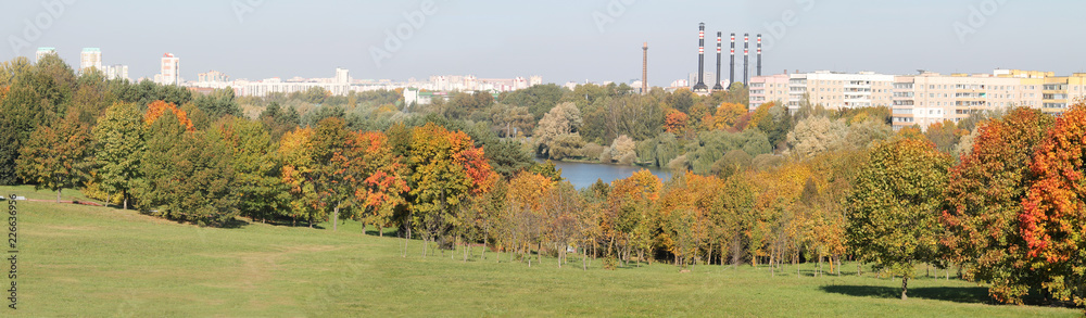Obraz premium Panoramic view of autumn park with deciduous trees that began to turn red and yellow on shore of lake. Minsk, Belarus