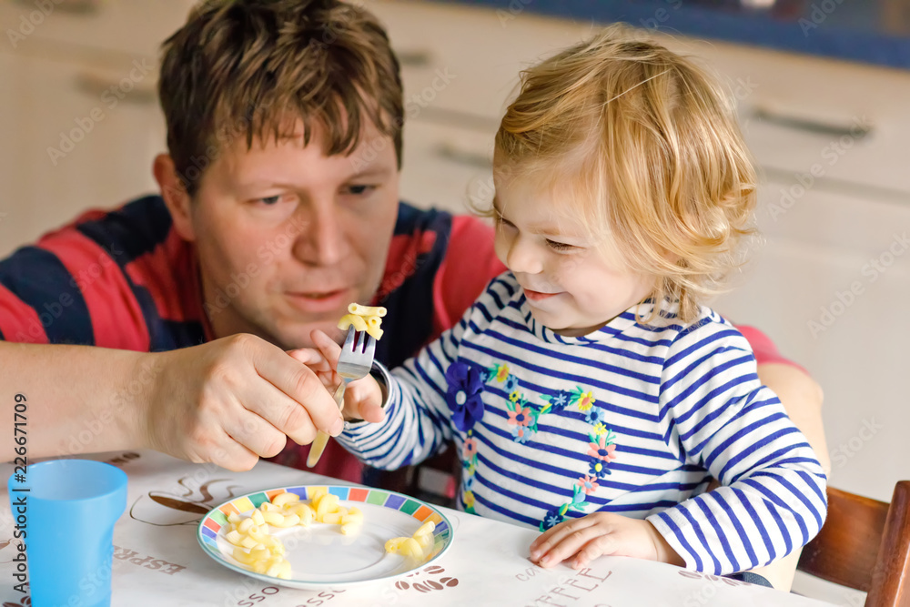Adorable baby girl eating noodle, pasta macaroni. Father helping daughter learning to use spoon. Cute healthy toddler child sitting in highchair, learning to eat by itself with dad in domestic kitchen Adorable baby girl eating noodle, pasta macaroni. Father helping daughter learning to use spoon. Cute healthy toddler child sitting in highchair, learning to eat by itself with dad in domestic kitchen