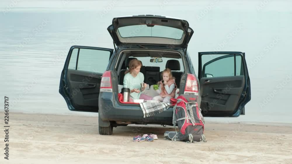 Beautiful young woman and her little daughter are sitting in the open trunk of a car on the bank of a river of the sea eating fruits and drinking tea from a thermos Beautiful young woman and her little daughter are sitting in the open trunk of a car on the bank of a river of the sea eating fruits and drinking tea from a thermos