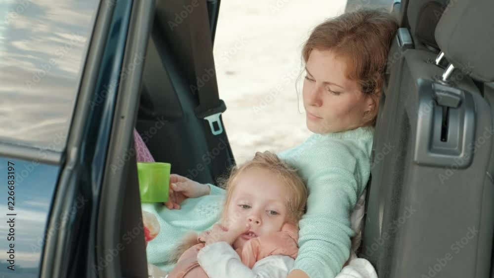 Beautiful young woman and her little daughter are sitting in the open trunk of a car on the bank of a river of the sea eating fruits and drinking tea from a thermos Beautiful young woman and her little daughter are sitting in the open trunk of a car on the bank of a river of the sea eating fruits and drinking tea from a thermos