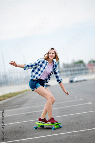 A beautiful smiling blond girl wearing checkered shirt, cap and denim shorts is longboarding while stretching out her hands. Sport and cool style.