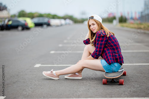 A beautiful blond girl wearing checkered shirt, cap and denim shorts is sitting on the longboard on the car park with a bold look. Sport and cool style.