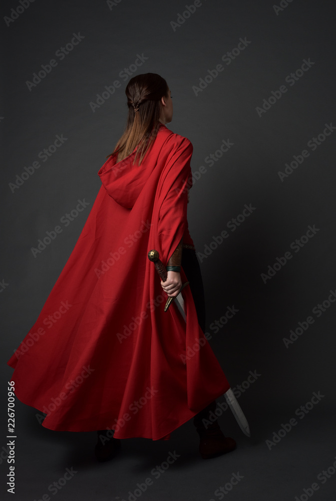 full length portrait of brunette girl wearing red medieval costume and cloak. standing pose with back to the camera, holding a sword on grey studio background. full length portrait of brunette girl wearing red medieval costume and cloak. standing pose with back to the camera, holding a sword on grey studio background.