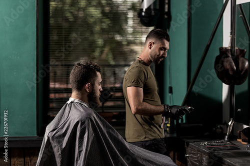 Brutal man sits at a barber shop in front the mirror. Barber takes the hairclipper