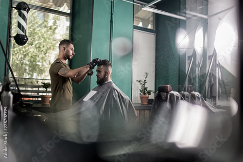 Young man with a beard sits in the chair at a barber shop. Barber dries mens hairs.