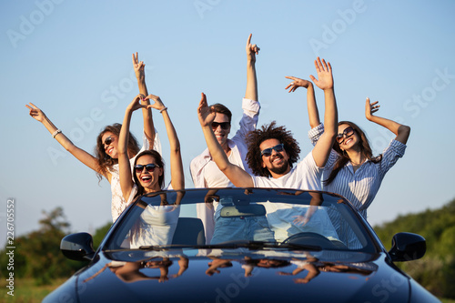 Happy young girls and guys in sunglasses are sitting in a black cabriolet on the road holding their hands up and smiling on a sunny day.