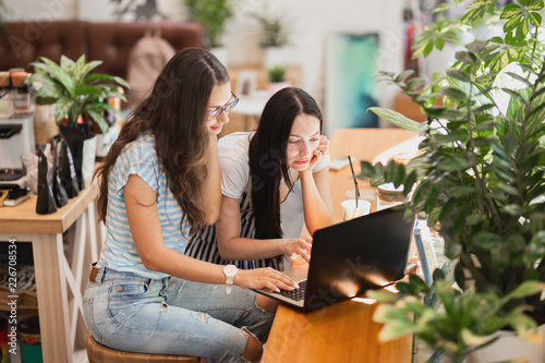 Two pretty slim girls with long dark hair,wearing casual style,sit at the table and look attentively at the laptop screen in a cozy coffee shop.