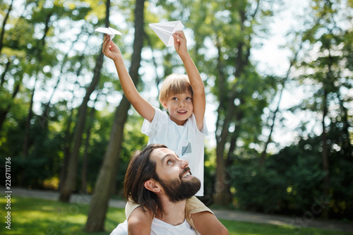 Father and his son walking in the forest. Boy dressed in a white t-shirt sitting on the shoulders of his father and keeps a paper airplane in his hand.