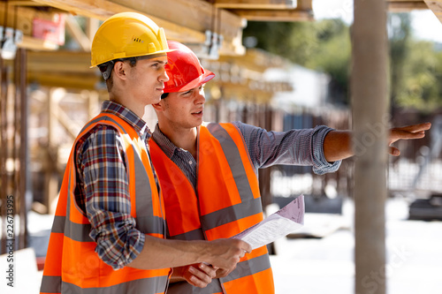 Two civil engineers dressed in orange work vests and helmets discuss the construction process on the building site near the wooden constructions and steel frames photo