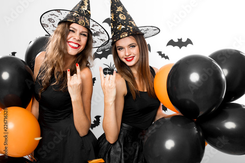 Two gorgious women in black dresses and witch`s hats hold black and orange balloons. Halloween photo