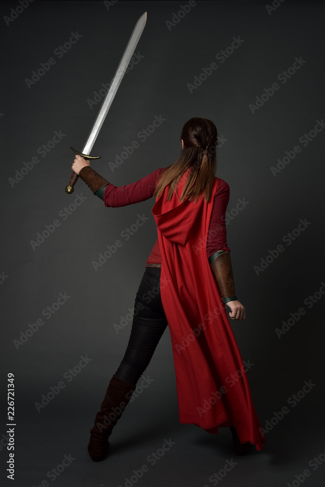 full length portrait of brunette girl wearing red medieval costume and cloak. standing pose with back to the camera, holding a sword on grey studio background. full length portrait of brunette girl wearing red medieval costume and cloak. standing pose with back to the camera, holding a sword on grey studio background.