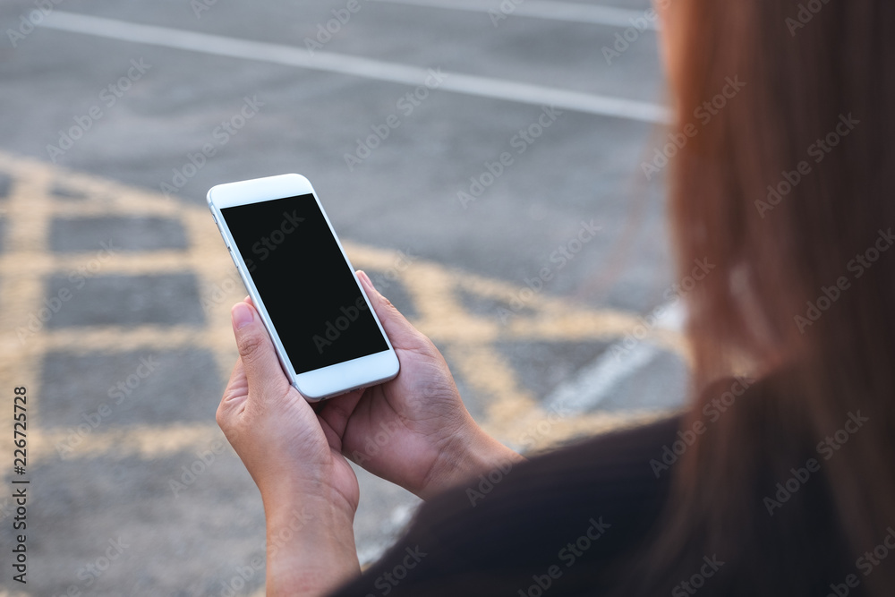 Mockup image of a woman holding white mobile phone with blank black screen with street background