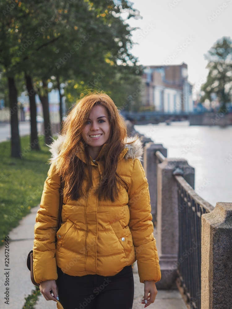 Brunette girl with long hair in a bright yellow jacket stands on the waterfront against the autumn trees