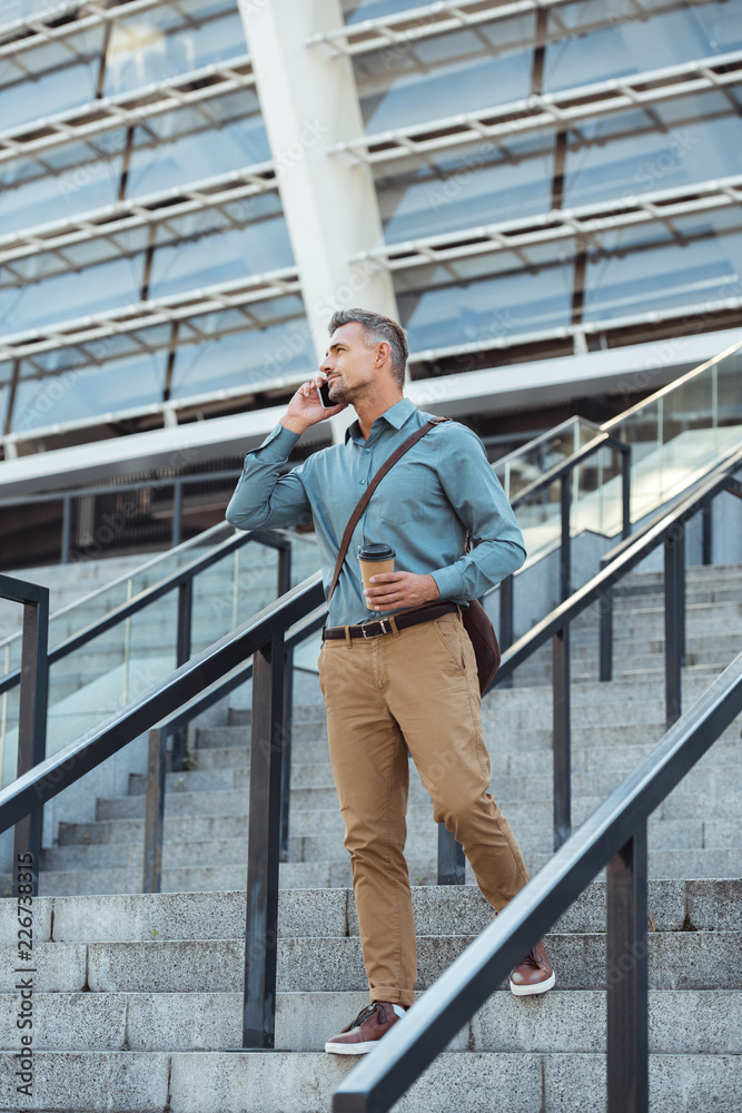 handsome man talking by smartphone and looking away while standing with coffee to go on stairs