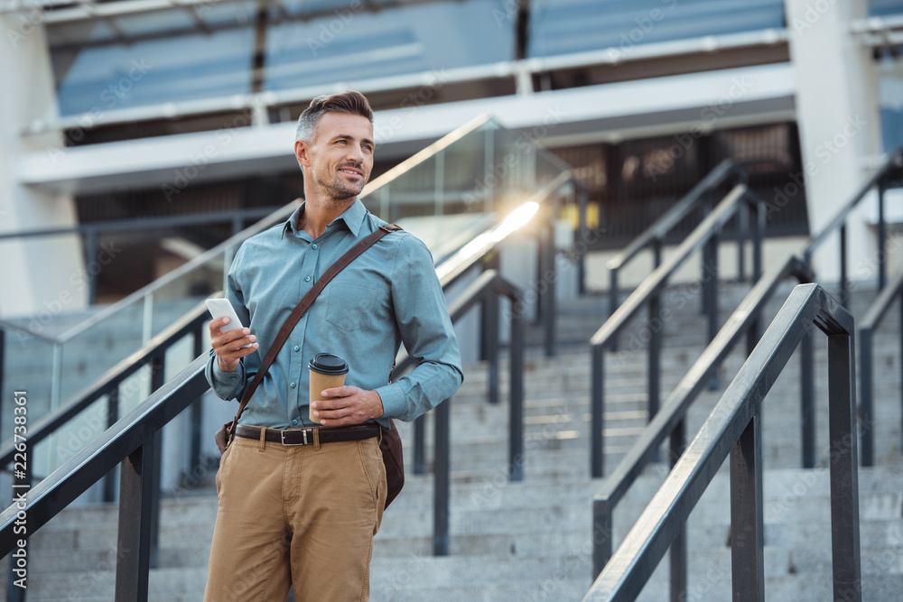 smiling middle aged businessman with coffee to go and smartphone looking away on stairs