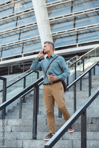 handsome man talking by smartphone and looking away while standing with coffee to go on stairs
