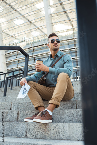 low angle view of man in sunglasses holding coffee to go and business newspaper while sitting on stairs