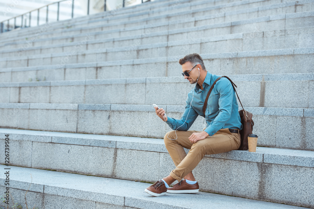 handsome stylish man in earphones and sunglasses sitting on stairs and using smartphone handsome stylish man in earphones and sunglasses sitting on stairs and using smartphone