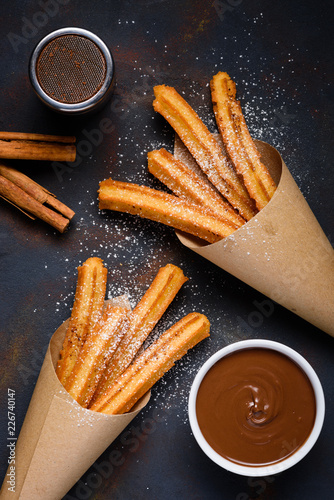 Churros with sugar powder with chocolate sauce dip and cinnamon sticks on dark background top view photo