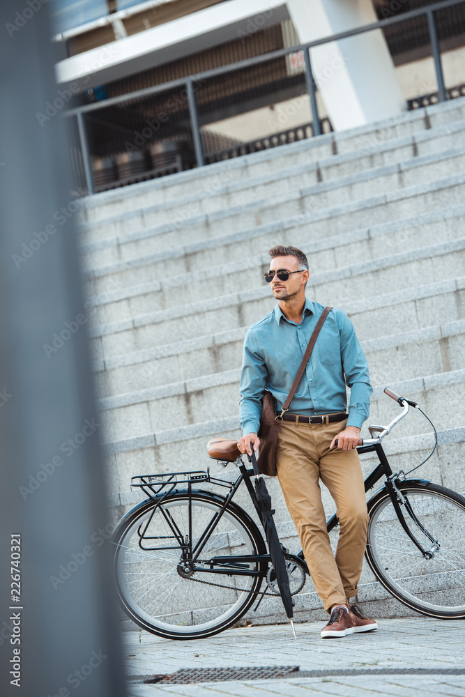handsome middle aged businessman in sunglasses holding umbrella while standing near bicycle on street handsome middle aged businessman in sunglasses holding umbrella while standing near bicycle on street