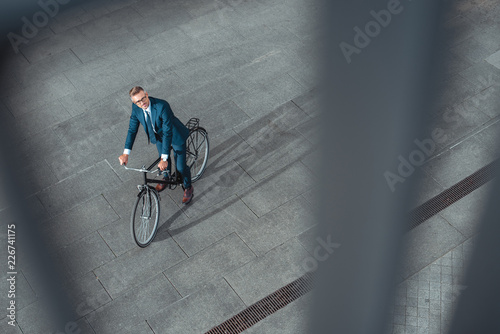 Fototapeta Naklejka Na Ścianę i Meble -  high angle view of handsome middle aged businessman in suit and eyeglasses riding bicycle