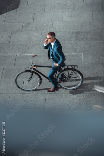 high angle view of businessman in formal wear talking by smartphone while standing with bicycle on street