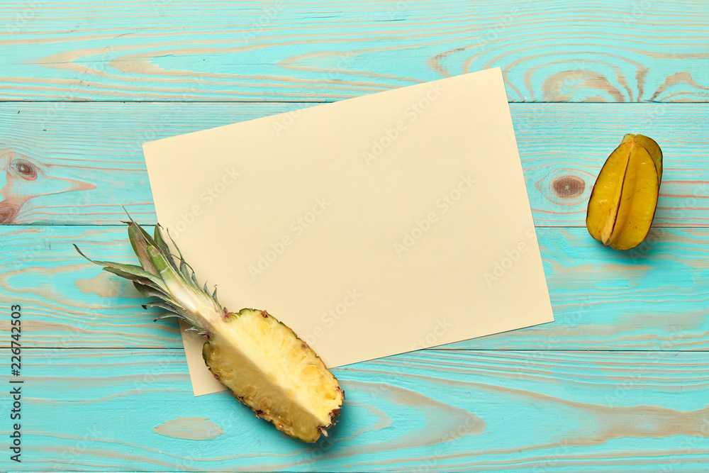 White cardboard with a slice of pineapple and a whole carambola on a blue wooden background with space for text. . Flat lay