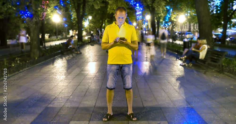 Timelapse of man standing still with smartphone on crowded walkway park boulevard at summer night. People moving fast. Concept of flowing time and technologies