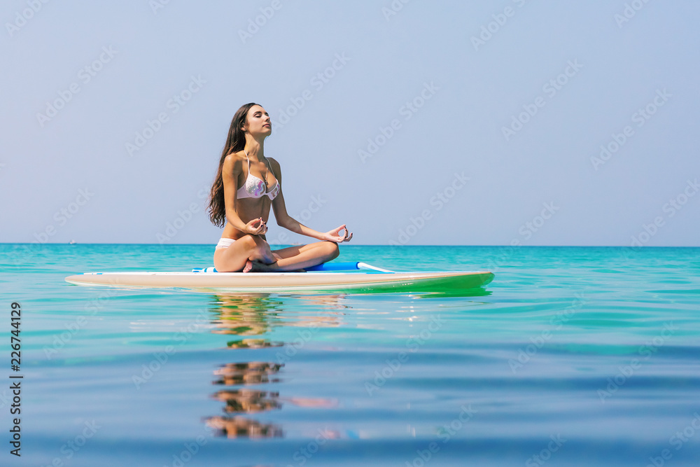 Young beautiful woman meditating in a sea at SUP paddleboarding. Healthy lifestyle. Girl in yoga pose relaxing in calm water.