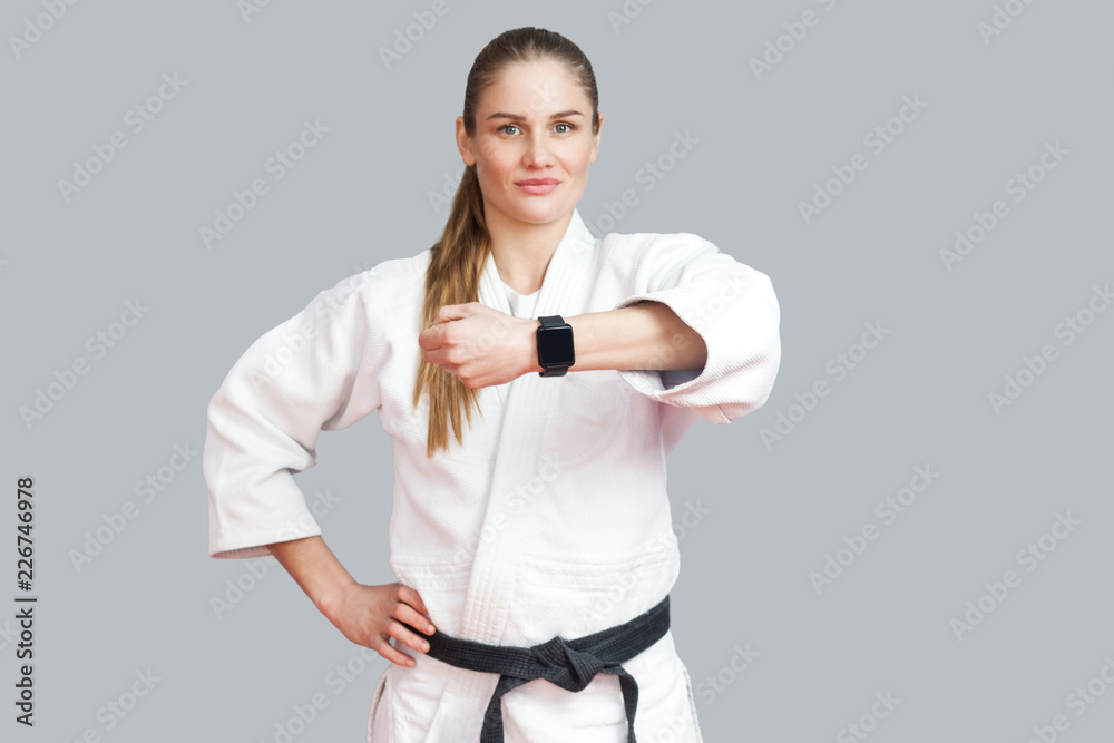Beautiful satisfied proud young blonde athletic woman posing and showing her smart watch, smile and looking at camera. Woman using fitness bracelet. Indoor studio shot, isolated on grey background Beautiful satisfied proud young blonde athletic woman posing and showing her smart watch, smile and looking at camera. Woman using fitness bracelet. Indoor studio shot, isolated on grey background