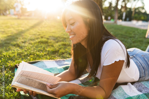 Smiling young asian girl outdoors in park reading book.