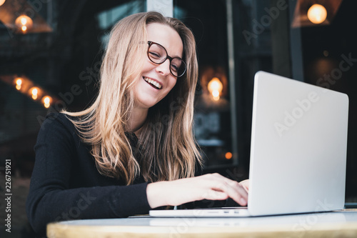 Portrait of a young beautiful smiling girl wearing eyeglasses, sitting at cafe, working on laptop. Concept of business comminication.   photo