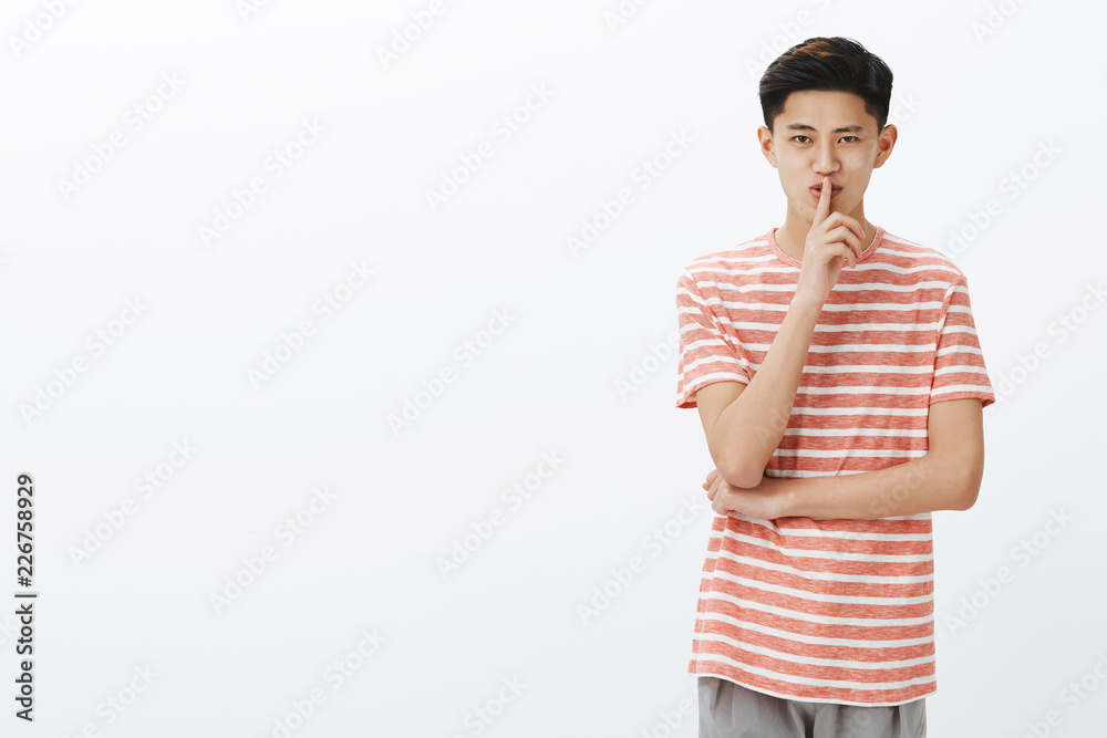 Portrait of charming mysterious young asian teenager with short hairstyle showing shush gesture and smiling having surprise or sharing secret, saying shh standing on right side of grey copy space Portrait of charming mysterious young asian teenager with short hairstyle showing shush gesture and smiling having surprise or sharing secret, saying shh standing on right side of grey copy space