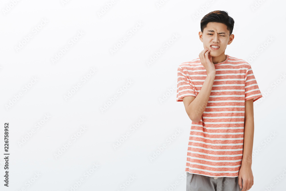 Uneasy attractive young asian male student having tooth decay touching cheek reacting on pain, having rotten teeth, suffering painful feeling while waiting in queue to dentist over grey wall Uneasy attractive young asian male student having tooth decay touching cheek reacting on pain, having rotten teeth, suffering painful feeling while waiting in queue to dentist over grey wall