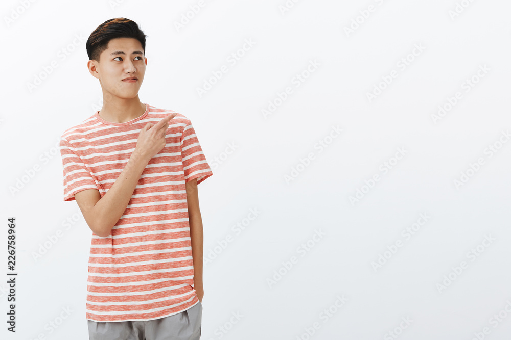 Portrait of curious nice young asian male model in striped t-shirt standing relaxed over grey wall with hand in pocket looking and pointing at upper right angle seeing interesting copy space Portrait of curious nice young asian male model in striped t-shirt standing relaxed over grey wall with hand in pocket looking and pointing at upper right angle seeing interesting copy space